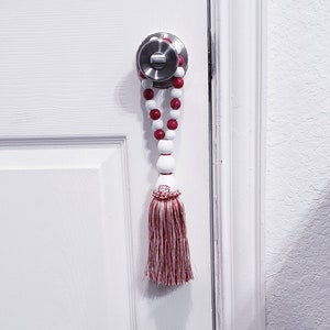 May include: A white door with a silver door knob and a red and white beaded tassel hanging from the knob.