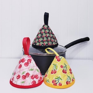 Puede incluir: Tres agarraderas cónicas con asas de lazo descansan sobre una olla negra. Las agarraderas presentan patrones de cerezas sobre fondos negros, rosas y amarillos. Los ribetes y asas de lazo rojos, amarillos y negros completan el diseño. Las agarraderas son para uso en la cocina.