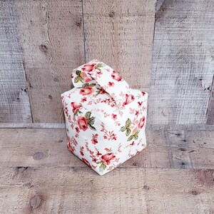 Könnte beinhalten: Ein weißer Türstopper mit einem Blumenmuster mit rosa Rosen und grünen Blättern. Der Türstopper ist würfelförmig und hat einen Griff oben.