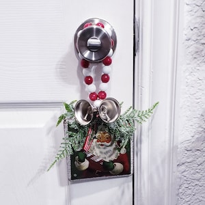 May include: A white door with a silver door knob and a Christmas decoration hanging from it. The decoration is a string of red and white beads with two silver bells and a framed picture of Santa Claus.