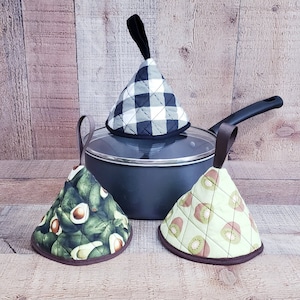 May include: Three pot holders in different patterns. One is black and white checkered, one has a green avocado pattern, and the other has a green kiwi pattern. The pot holders are on a wooden surface.