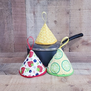 Puede incluir: Tres agarraderas de olla en forma de cono. Las agarraderas están hechas de tela y tienen diferentes patrones. Una agarradera tiene un patrón de panal de abeja amarillo, otra tiene un patrón de fresa roja y la tercera tiene un patrón de lima verde.