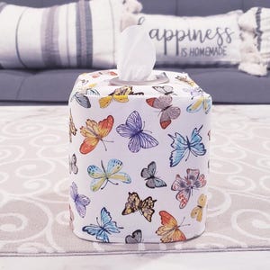 Pu&ograve; includere: Un copri scatola di fazzoletti bianco con un motivo a farfalla. Il copri scatola presenta una variet&agrave; di farfalle colorate su uno sfondo bianco. Il testo "Happiness is Homemade" &egrave; stampato sulla parte superiore del copri scatola.