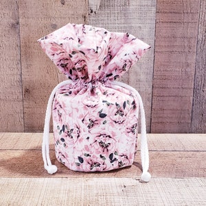 K&ouml;nnte beinhalten: Ein Kordelzugbeutel mit einem floralen Muster aus rosa Rosen und gr&uuml;nen Bl&auml;ttern. Der Beutel ist mit einer wei&szlig;en Kordel gebunden. Der Hintergrund ist eine Holzoberfl&auml;che.