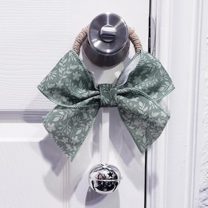 May include: A green floral patterned bow with a silver jingle bell hanging from it. The bow is tied around a door knob.