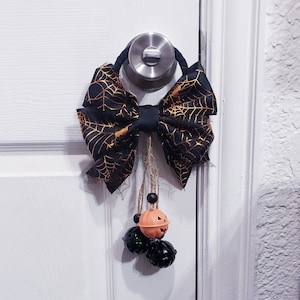 May include: A black and gold bow with a spiderweb pattern hangs from a door knob.  Three small bells hang from the bow, one with a jack-o-lantern face.