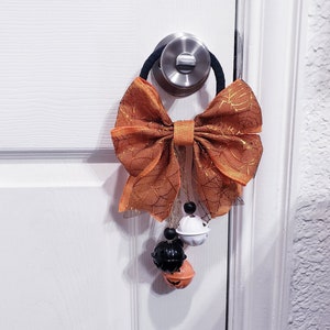 May include: A Halloween door decoration with an orange bow and three jingle bells. The bells are decorated with a pumpkin, a ghost, and a black cat.