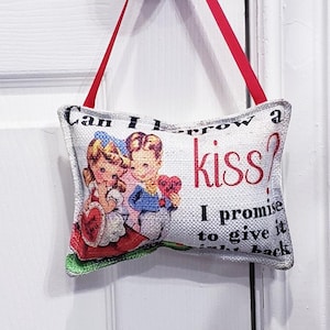 May include: A rectangular decorative pillow with a vintage illustration of a couple and the text "Can I borrow a kiss? I promise to give it right back." The pillow is suspended from a red ribbon.