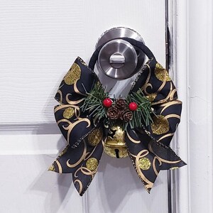 Puede incluir: Un lazo negro y dorado con una campanita y ramitas de pino cuelga de un pomo de puerta blanco.