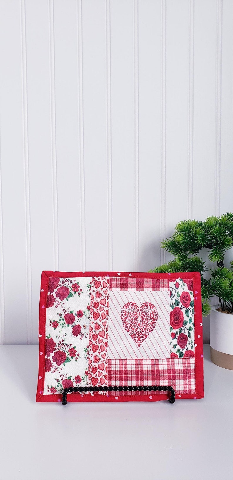 Puede incluir: Un posavasos acolchado rojo y blanco con un dise&ntilde;o de coraz&oacute;n. El posavasos est&aacute; hecho de tela con un patr&oacute;n de patchwork de corazones, flores y cuadros. Se muestra sobre una superficie blanca con una peque&ntilde;a planta en maceta en el fondo.