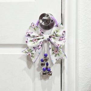 Puede incluir: Un lazo morado y blanco con estampado de mariposas y una cadena de cascabeles plateados colgando de él. El lazo está atado alrededor de un pomo de puerta.