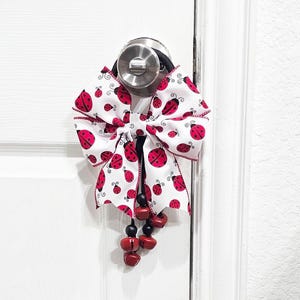 Könnte beinhalten: Eine weiße Schleife mit roten Marienkäfern ist um einen Türknauf gebunden. Eine Schnur mit roten Glöckchen hängt von der Schleife.