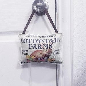 May include: A white doorstop with a brown strap hanging from a door knob. The doorstop is a small pillow with the text "Coffee & Bakery Cottontail Farms" and a picture of a rabbit and eggs. The text "Fresh Made" and "Open Daily" is also on the pillow.