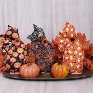 May include: Three Halloween-themed fabric figures with pumpkin, spiderweb, and ghost patterns. They are displayed with two ceramic pumpkins on a wooden tray, surrounded by fall leaves. The figures are on wooden bases.