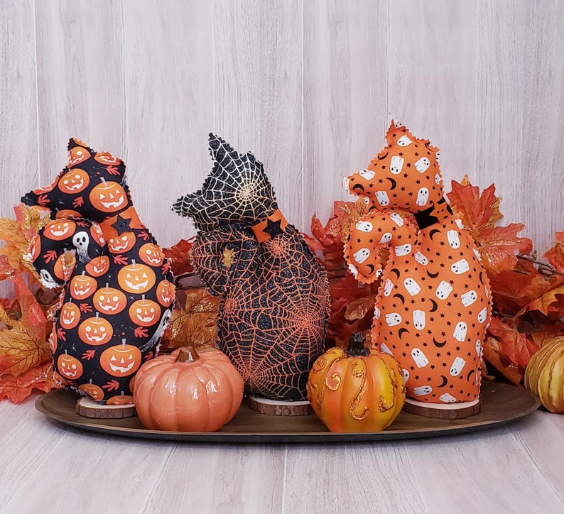May include: Three Halloween-themed fabric figures with pumpkin, spiderweb, and ghost patterns. They are displayed with two ceramic pumpkins on a wooden tray, surrounded by fall leaves. The figures are on wooden bases.