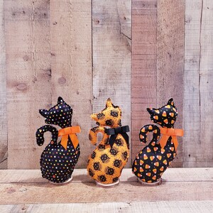 May include: Three black felt Halloween cat figurines with orange bows. The cats are standing on wooden bases. The cats are decorated with different patterns: black and white polka dots, black cats on an orange background, and candy corn.