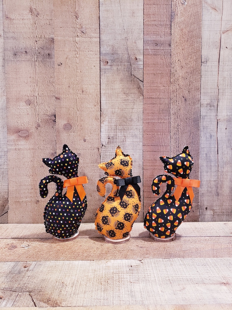 May include: Three black felt Halloween cat figurines with orange bows. The cats are standing on wooden bases. The cats are decorated with different patterns: black and white polka dots, black cats on an orange background, and candy corn.