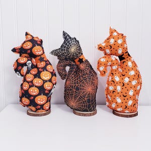 May include: Three Halloween-themed fabric figures on wooden bases. The figures have witch-like shapes and are decorated with pumpkin, spiderweb, and ghost patterns in orange, black, and white. The figures are displayed on a white surface.