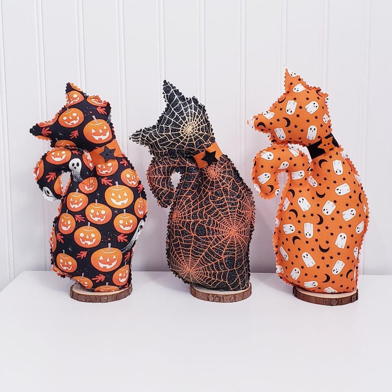 May include: Three Halloween-themed fabric figures on wooden bases. The figures have witch-like shapes and are decorated with pumpkin, spiderweb, and ghost patterns in orange, black, and white. The figures are displayed on a white surface.