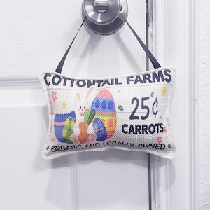 May include: A white door with a silver door knob. A small burlap sign hangs from the knob. The sign says "Cottontail Farms 25¢ Carrots Organic and Locally Owned". The sign has a cartoon bunny holding a carrot and an Easter egg.