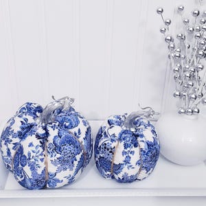 May include: Two decorative pumpkins with a blue and white floral pattern, silver stems, and twine accents. They sit on a white tray, alongside a white vase with silver decorative stems. The pumpkins are a fall-themed decorative item.