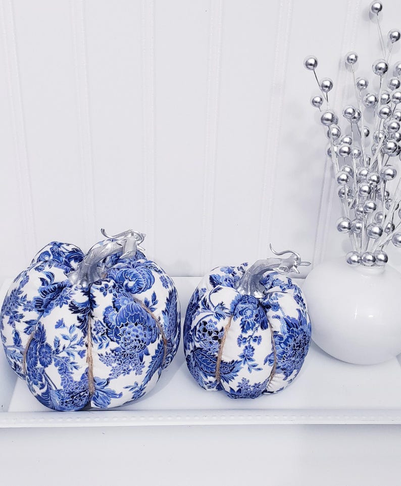 May include: Two decorative pumpkins with a blue and white floral pattern, silver stems, and twine accents. They sit on a white tray, alongside a white vase with silver decorative stems. The pumpkins are a fall-themed decorative item.