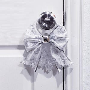May include: A white door with a silver doorknob is adorned with a decorative bow. The bow is white with silver snowflake patterns and a silver trim. A silver bell is in the center of the bow.