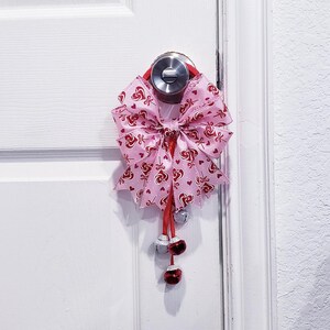 May include: A pink bow with a pattern of red hearts and lollipops hangs on a white door. The bow is adorned with silver and red jingle bells. The door knob is silver.