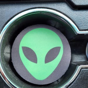 May include: Green alien face printed on a black and gray car cup holder.