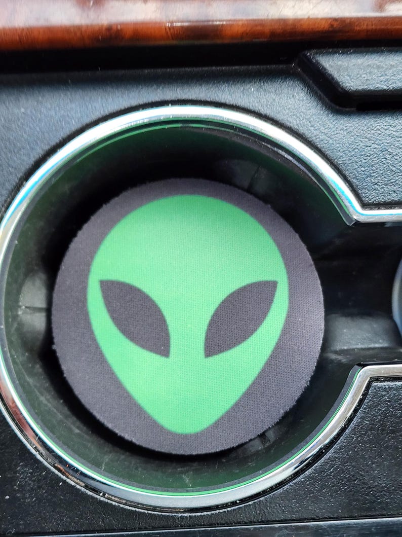 May include: Green alien face printed on a black and gray car cup holder.