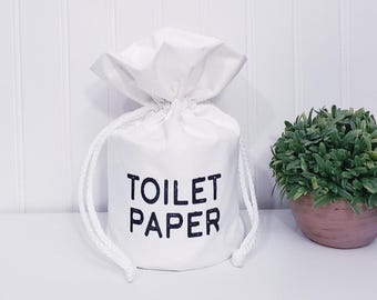 May include: White fabric toilet paper holder with a drawstring closure. The holder has black embroidered text that reads "TOILET PAPER".