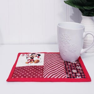 Könnte beinhalten: Eine weiße Keramiktasse mit Schneeflockenmustern steht auf einem weihnachtlichen Patchwork-Untersetzer. Der Untersetzer zeigt eine Mickey Mouse-Grafik mit einer Zuckerstange, rot-weißen Streifen und einem roten Rand.