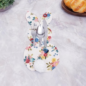 Puede incluir: Porta cubiertos con forma de conejito con un estampado floral en rosa, azul y amarillo. El soporte tiene una cola de pompón blanco y contiene un tenedor, una cuchara y un cuchillo. El fondo es una superficie de mármol.