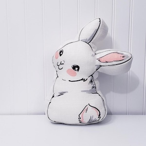 May include: A white plush bunny with pink ears and a black embroidered face. The bunny is sitting on a white surface.