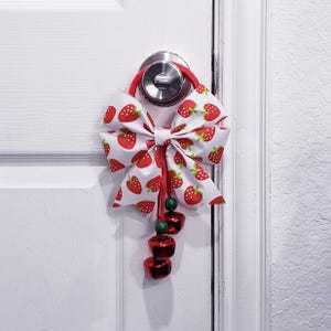 May include: A white fabric bow with a red strawberry pattern hangs from a door handle. The bow is accented with red ribbon and green beads, and three red bells dangle below. The door is white.