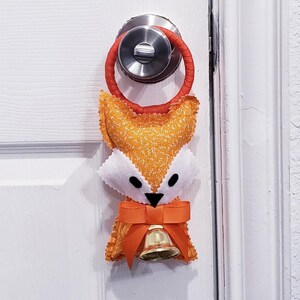 Puede incluir: Un colgador de puerta de zorro naranja y blanco hecho a mano con un lazo rojo, un lazo y una pequeña campana dorada. El zorro tiene ojos negros y está sujeto a un pomo de puerta.