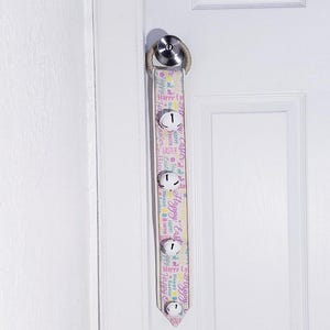 May include: A white door with a decorative Easter door hanger. The hanger is made of fabric with a pink, yellow, and blue pattern that says "Happy Easter" and has four small bells.