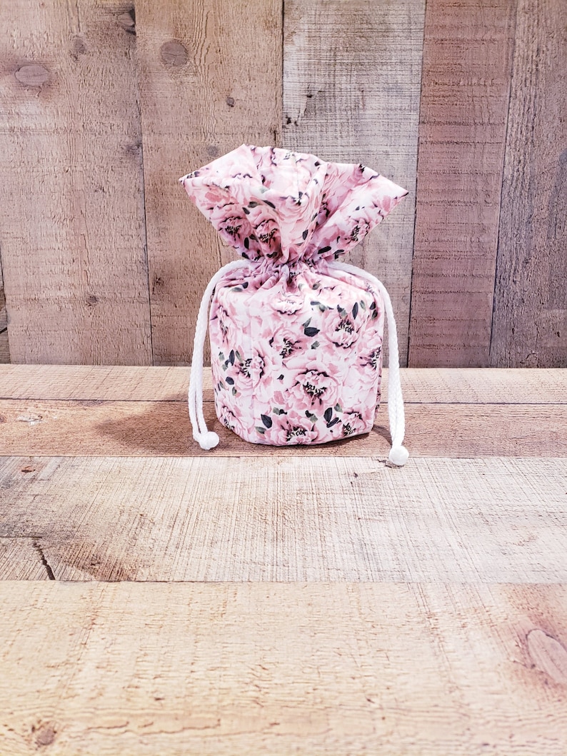 K&ouml;nnte beinhalten: Ein Kordelzugbeutel mit einem floralen Muster aus rosa Rosen und gr&uuml;nen Bl&auml;ttern. Der Beutel ist mit einer wei&szlig;en Kordel gebunden. Der Hintergrund ist eine Holzoberfl&auml;che.