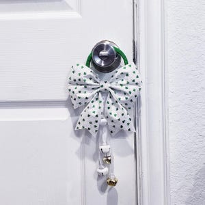 Puede incluir: Un lazo de tela blanca con un estampado de tréboles verdes, colgado de una puerta. El lazo está sujeto a un lazo verde alrededor del pomo de la puerta. Cintas blancas con pequeñas campanas cuelgan del lazo, con detalles dorados y plateados.