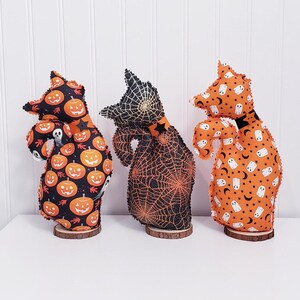 May include: Three Halloween-themed fabric figures on wooden bases. The figures have a cat-like shape and are decorated with Halloween patterns, including pumpkins, spiderwebs, ghosts, and stars. The color scheme is primarily orange, black, and white.