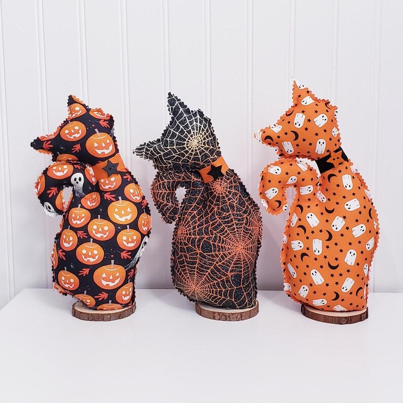 May include: Three Halloween-themed fabric figures on wooden bases. The figures have a cat-like shape and are decorated with Halloween patterns, including pumpkins, spiderwebs, ghosts, and stars. The color scheme is primarily orange, black, and white.