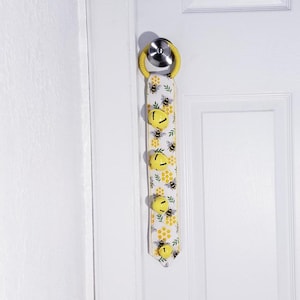 May include: A yellow and white door hanger with a bee and honeycomb pattern. The hanger has five yellow bells attached to it. The hanger is attached to a white door with a silver door knob.