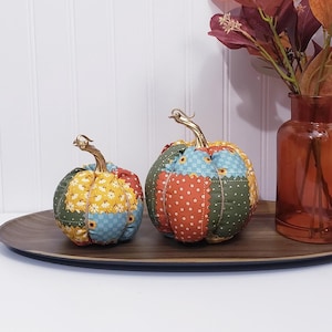 May include: Two stuffed fabric pumpkins featuring a patchwork design of various autumn-colored patterns including sunflowers, checkered prints, and dots. Each pumpkin is topped with a metallic gold stem and tied with thin twine.
