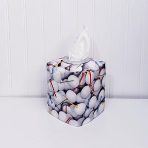 May include: A square tissue box cover featuring a golf ball pattern with golf tees in various colors. The tissue box has a silver rim and white tissues are visible. The design is printed on a fabric cover.