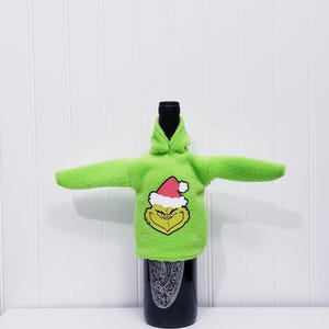 May include: A green bottle cover shaped like a hoodie, featuring the Grinch's face wearing a Santa hat. The cover is on a black bottle with a decorative label. The background is a white wall.