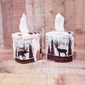 Op de afbeelding: Twee vierkante tissueboxhoezen met een bosmotief. Een hoes heeft een eland en de tekst "Moose" en de andere hoes heeft een hert en de tekst "Deer".