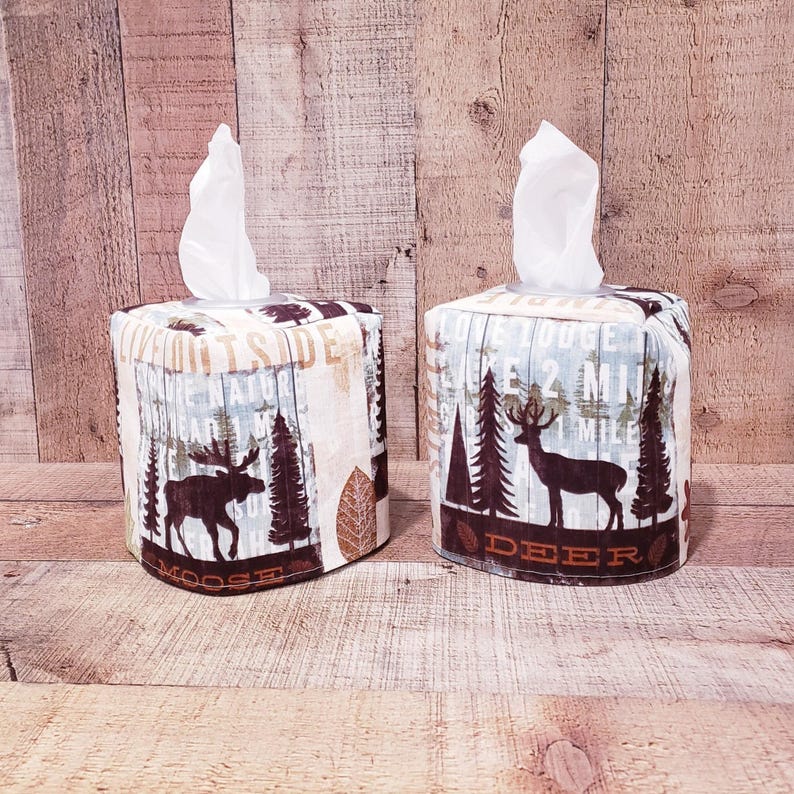 Op de afbeelding: Twee vierkante tissueboxhoezen met een bosmotief. Een hoes heeft een eland en de tekst "Moose" en de andere hoes heeft een hert en de tekst "Deer".
