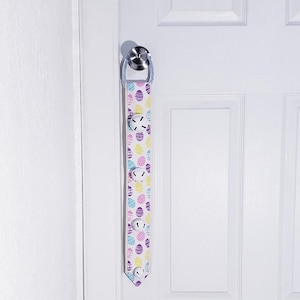 May include: A white door with a door handle and a fabric Easter door hanger with a white background and a pattern of colourful Easter eggs. The hanger has four white bells attached to it.