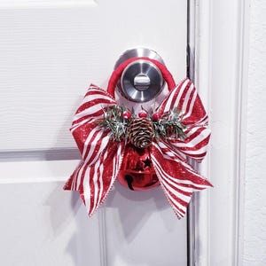 May include: A festive red and white striped bow and bell door hanger. The bow is accented with a pinecone, berries, and greenery. The red bell has a star cutout and hangs below the bow.