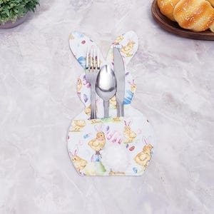 Puede incluir: Porta cubiertos con temática de Pascua en forma de conejito, con tenedor, cuchara y cuchillo. El soporte es azul claro con un estampado de pollitos, conejitos y huevos de Pascua. Lleva una cola de pompón blanco.